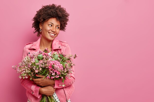 Studio shot of glad woman holds big bouquet of flowers celebrates spring holiday smiles gladfully looks away poses against pink wall with copy space for your promotion