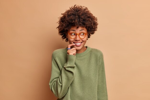 Studio shot of glad woman looks with curious happy gaze aside keeps finger near lips smiles broadly dressed casually isolated over beige wall listnes interesting rumors