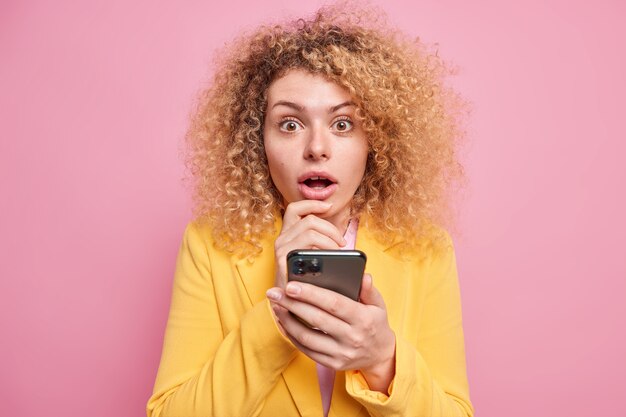 Студийный снимок красивой европейской женщины с кудрявыми волосами, получившей невероятные новости, держит смартфон, отправляет текстовые сообщения, держит рот открытым