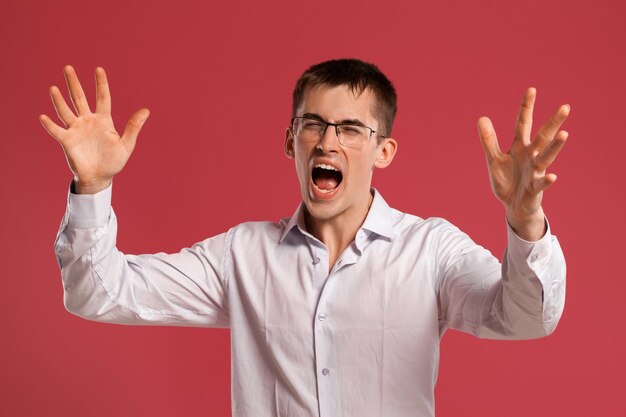 Studio shot of a good-looking young man in a classic white shirt, black watches and glasses screaming while posing over a pink background. Stylish haircut. Sincere emotions concept. Copy space.