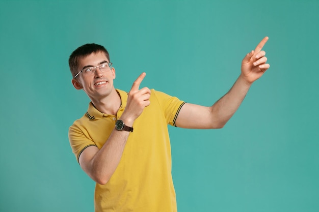 Studio shot of a good-looking young man in a yellow casual t-shirt, glasses and black watches pointing at something while posing over a blue background. Stylish haircut. Sincere emotions concept. Copy