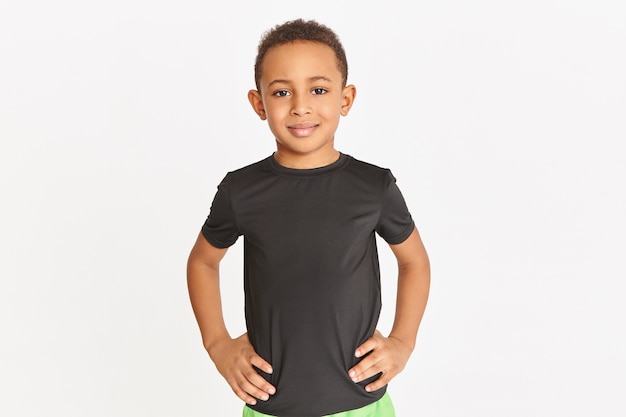 Studio shot of handsome athletic dark skinned little boy posing isolated in black t-shirt keeping hands on his waist, training indoors.