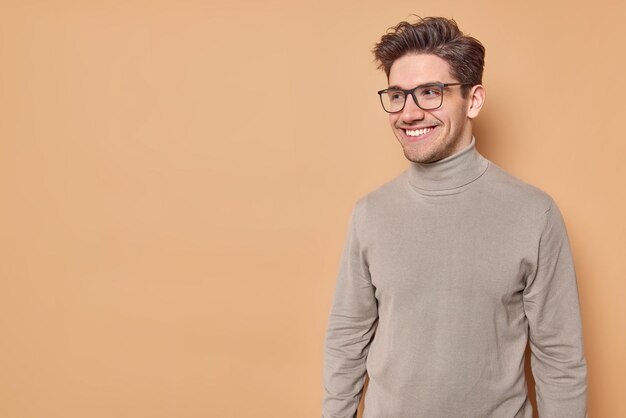 ハンサムなヨーロッパ人男性のスタジオショットは、どこかに焦点を当てた良い気分で積極的に微笑んでいますあなたのプロモーションのためのコピースペースでベージュの背景に眼鏡とタートルネックのポーズを着ています