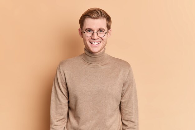 Studio shot of handsome teenage boy smiles pleasantly has happy talk wears round transparent glasses and poloneck 