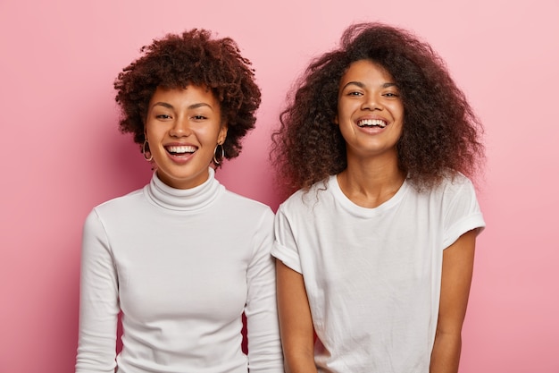 Studio shot of happy Afro American female sisters enjoy good moment, wear white casual clothes, smile broadly, have fun together during free time, isolated over pink wall.