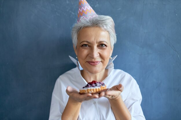 Studio shot of happy beauitful middle aged female wearing conical hat celebrating birthday, posing isolated with cake in her hands, offering you to have bite. Selective focus on woman's face
