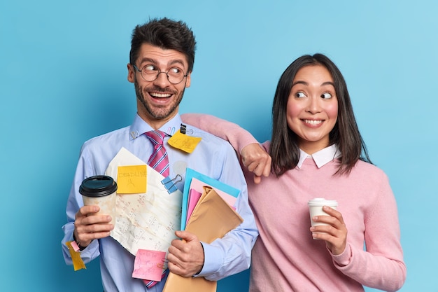 Studio shot of happy classmates discuss school project ideas stand glad drink take away coffee look cheerfully at each other. Glad nerd man holds folders covered with stickers on formal shirt