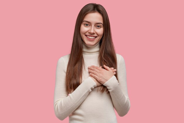 Студийный снимок счастливой милой молодой женщины, которая складывает руки на груди, выражает благодарность, ругается или что-то обещает