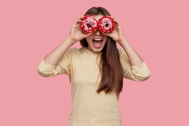 Studio shot of happy dark haired woman covers eyes with two red donuts, being in high spirit, wears yellow clothes