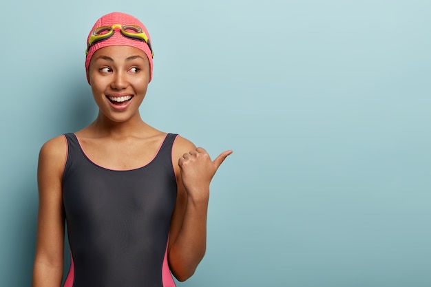 Студийный снимок счастливой темнокожей женщины с очаровательной улыбкой, в купальных костюмах, показывает пальцем в сторону