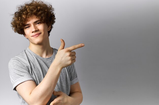 Studio shot of happy energetic young Caucasian male indicating copy space gray wall pointing index finger away and smiling at camera, having cheerful positive facial expression