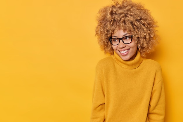 Studio shot of happy sincere young european woman with curly hair concentrated away gladfully wears optical glasses and casual jumper isolated over yellow background. human emotions concept
