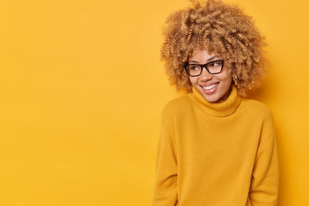 Studio shot of happy sincere young European woman with curly hair concentrated away gladfully wears optical glasses and casual jumper isolated over yellow background. Human emotions concept.