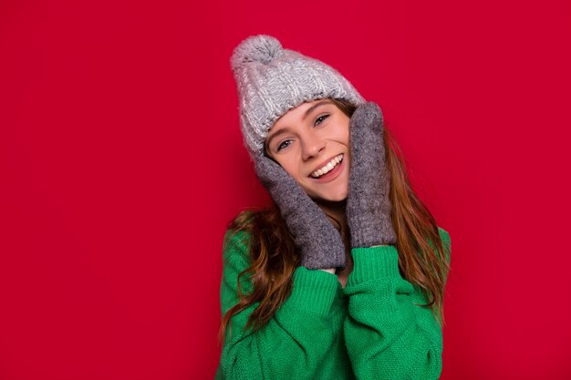 Studio shot of happy smiling girl with adorable smile and blue eyes touching her face dressed winter cap and mittens on isolated red background