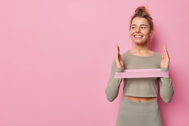 Free photo studio shot of happy sporty european woman stretches elastic band does exercises for hands dressed in activewear looks away poses against pink background blank copy space for your promotion.