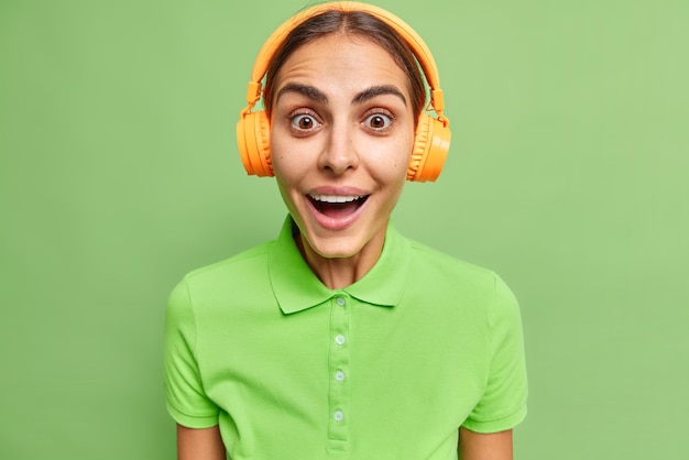 Studio shot of happy surprised woman looks with great interest wears casual t shirt listens music via wireless headphones reacts on amazing news isolated over vivid green background. Female meloman