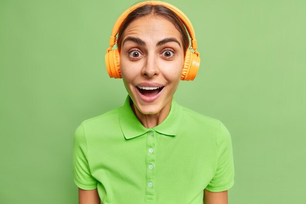 큰 관심을 가지고 있는 행복한 놀란 여성의 스튜디오 샷은 캐주얼한 티셔츠를 입고 무선 헤드폰을 통해 음악을 듣고 선명한 녹색 배경에서 격리된 놀라운 뉴스에 반응합니다. 여성 멜로맨