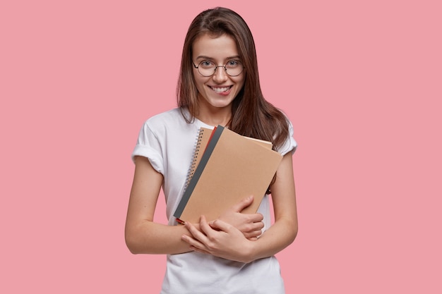 Studio shot of happy teenage girl bites lower lip, carries notepads, looks joyfully , wears white t shirt, round spectacles