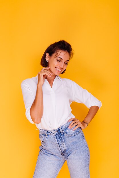 Studio shot of happy woman short hair wearing white shirt and denim pants posing on yellow wall