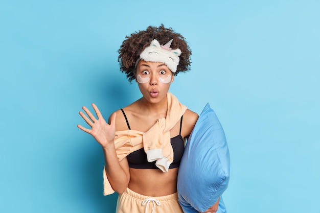 Studio shot of impressed curly haired woman raises palm and stares at camera prepares for sleep holds soft pillow