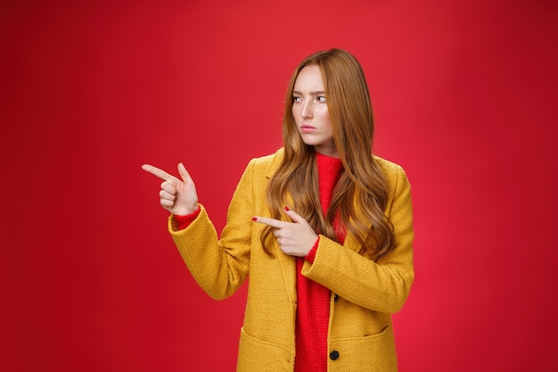 Studio shot of intense suspicious and displeased redhead female in yellow coat pointing and looking ...