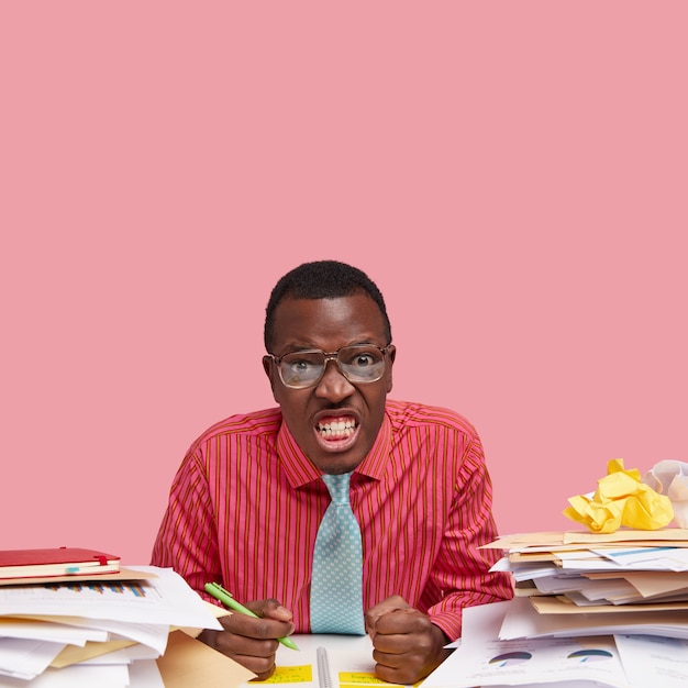 Studio shot of irritated dark skinned man clenches teeth, keeps hands in fists, dressed in formal clothes