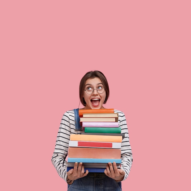 Studio shot of joyful European woman wears round spectacles, holds many books