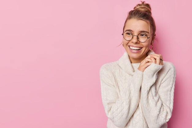 Studio shot of lovely cheerful woman keeps hands together smiles broadly looks away feels satisfied wears knitted sweater isolated over pink background empty space for your promotional content