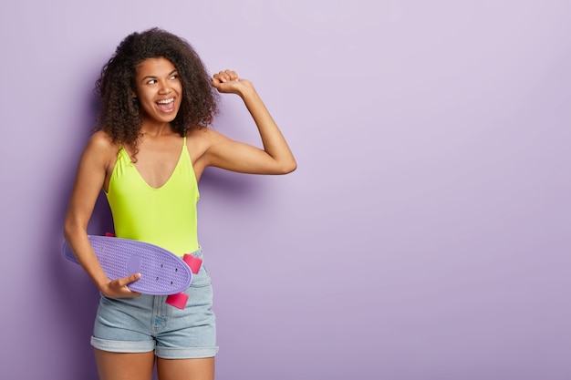 Studio shot of lovely curly haired woman poses with skateboard, takes break after active ride, has dark skin