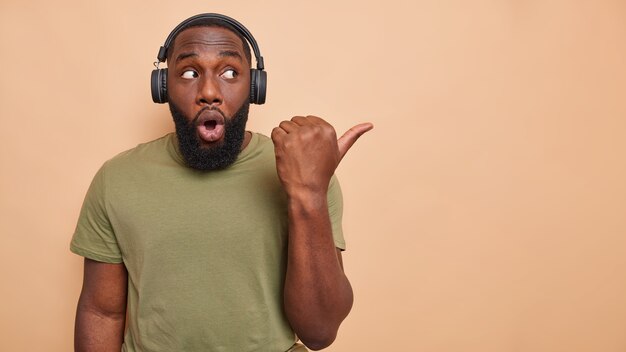 Studio shot of man with thick beard points thumb away on blank space listens music via wireless headphones dressed in casual t shirt isolated over beige wall