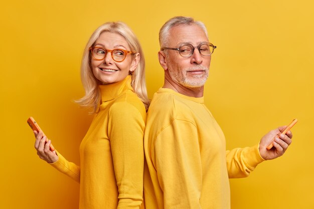 Studio shot of mature male and female models stand back to each other with modern gadgets in hands surf internet chat in social networks isolated over yellow wall