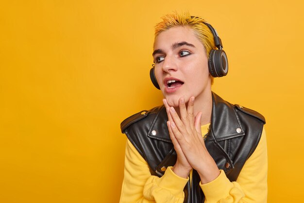 Studio shot of millennial girl keeps hands on chin concentrated away with curious expression listens music in wireless stereo headphones isolated on yellow blank copy space away