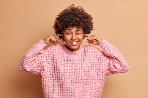 無料写真 美しい女性のスタジオショットは、大きな音を避けるために耳を塞ぎ、大きな音を止めるように求めますベージュの壁に隔離されたニットのセーターを着ています