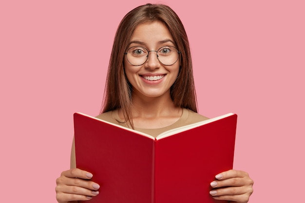 Бесплатное фото Студийный снимок довольной жизнерадостной женщины с мирным спокойным счастливым выражением лица держит раскрытый учебник перед