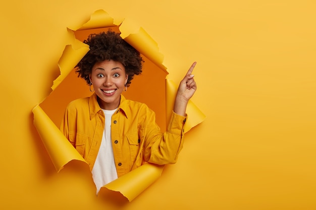 無料写真 ポジティブなアフリカ系アメリカ人女性のスタジオショットは、上のスペースをコピーするために指を指して、良い情報に興奮し、心地よく微笑んで、黄色いジャケットを着て、破れた紙の穴に立っています。