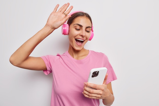 Studio shot of optimistic carefree woman dances with rhythm of music wears stereo headphones enjoys favorite song drressed in casual pink t shirt isolated over white background Hobby concept