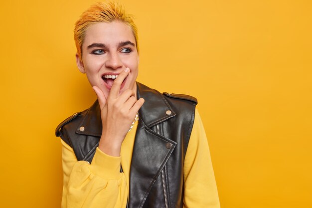 Studio shot of optimistic hipster girl laughs positively looks away keeps hand on mouth wears black leather vest casual jumper isolated over vivid yellow wall