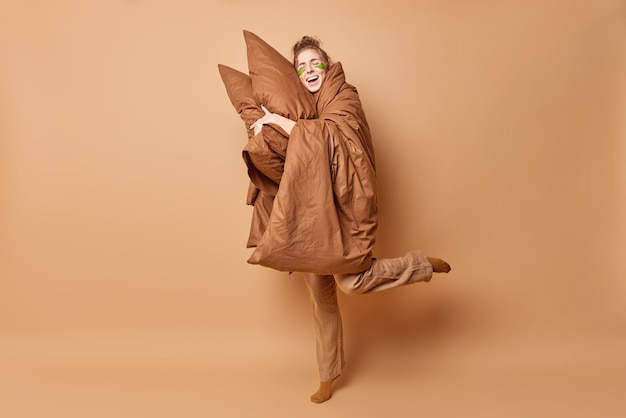 Studio shot of optimistic young woman leans on soft pillows takes nap wears pajama covered with blanket stands on one leg has good rest isolated over brown background. Sleeping time concept.