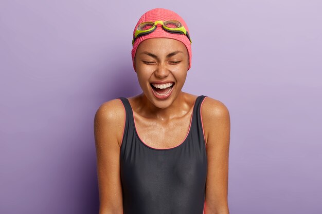 Studio shot of overjoyed dark skinned woman closes eyes, dressed in swimming costume, goggles on head, spends free time in sport center, has wet body, happy to finish race. Diving and exercising