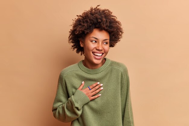 Studio shot of overjoyed woman laughs out loudly as hears funny story keeps hand on chest and looks on right smiles broadly dressed in casual jumper isolated over beige wall