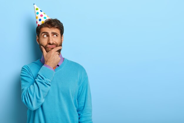 Студийный снимок задумчивого парня в шляпе на день рождения, позирующего в синем свитере