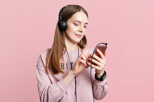 Studio shot of pleasant looking femal reads news online on cellphone or watches video in headphones