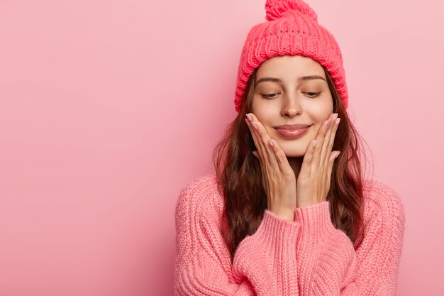 Studio shot of pleasant looking female model touches chin with both palms, has eyes closed,  has dark hair, dressed in winter outfit, isolated on pink wall, blank space