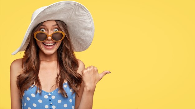 Studio shot of pleasant looking gorgeous young lady in trendy white hat, shows blank space or direction