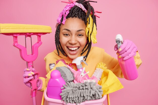 Studio shot of positive dark skinned woman cleans apartment smiles gladfully holds mop and cleaning detergent