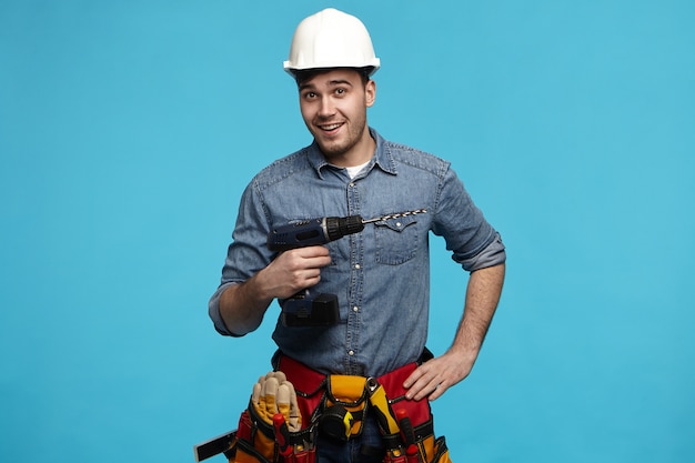 Студийный снимок позитивного дружелюбного молодого конструктора, несущего дрель и ремень со всеми инструментами, ремонтирующими для клиента