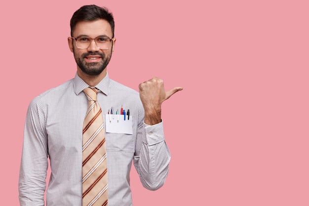 Free photo studio shot of positive unshaven european young man points with thumb aside, demonstrates result of his diligent work, feels proud