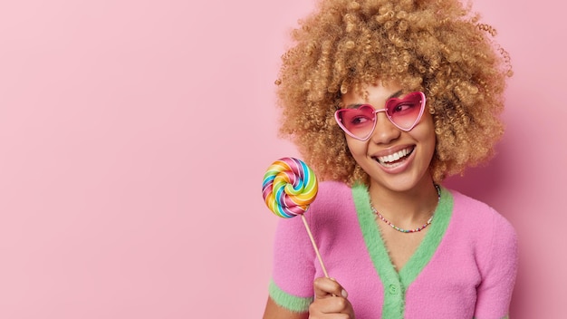 곱슬머리 미소를 가진 긍정적인 여성의 스튜디오 사진은 하트 모양의 선글라스를 광범위하게 착용하고 티셔츠에는 광고 콘텐츠를 위한 분홍색 배경 빈 공간 위에 격리된 다채로운 사탕이 들어 있습니다
