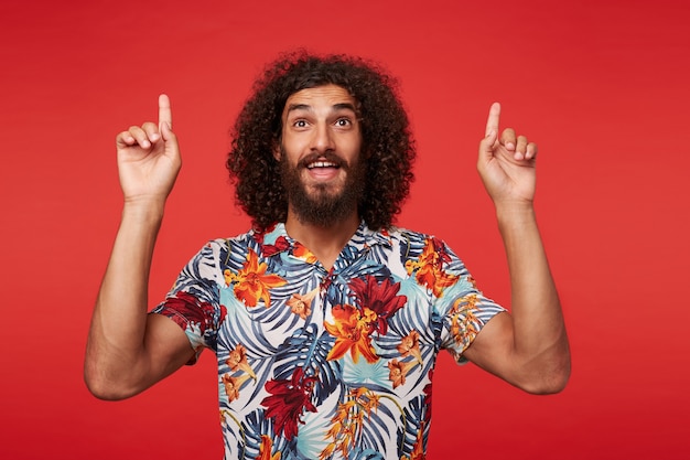 Studio shot of positive young brunette curly man with beard showing up with index fingers while posing against red background in multi-colored flowered shirt