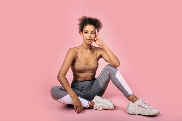 Studio shot of pretty black woman with african hairstyle in stylish sportwear sitting on pink background.